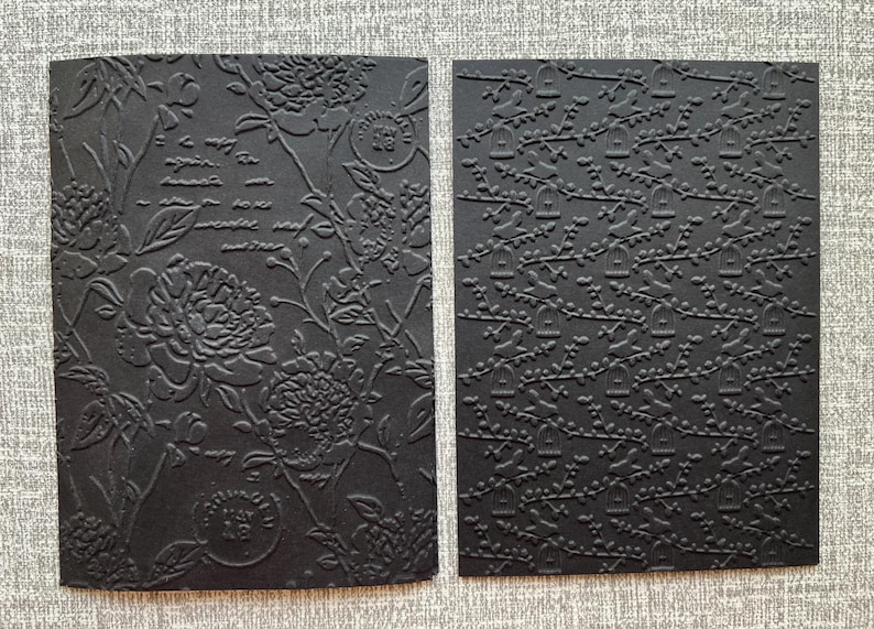 K&ouml;nnte beinhalten: Zwei schwarze, gepr&auml;gte Papierb&ouml;gen. Einer zeigt florale und Schriftmuster, der andere ein Muster aus Vogelk&auml;figen auf &Auml;sten. Das Papier hat ein strukturiertes Reliefdesign, ideal f&uuml;r Bastelarbeiten oder Schreibwaren.