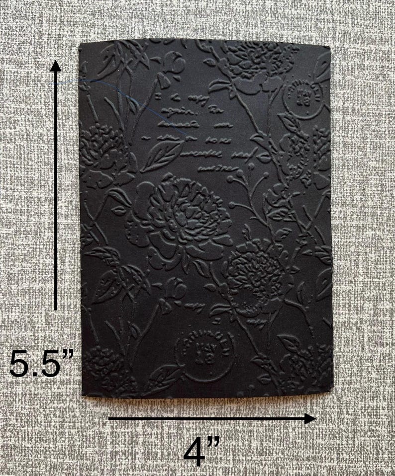 K&ouml;nnte beinhalten: Ein schwarzes, rechteckiges, gepr&auml;gtes Papier mit floralem Design und Schrift. Das Papier misst 14 cm x 10 cm. Das florale Muster umfasst gro&szlig;e, pfingstrosenartige Bl&uuml;ten und Bl&auml;tter. Die Schrift ist teilweise sichtbar.