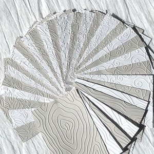 May include: A fan of 20 different embossed paper sheets in various shades of gray. Each sheet has a unique pattern, including swirls, leaves, and lines.