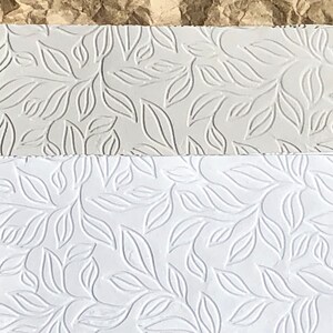 May include: Two sheets of white paper with an embossed leaf pattern. The top sheet is a light gray color and the bottom sheet is a bright white.
