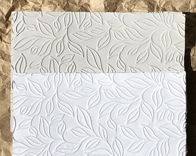 May include: Two sheets of white paper with an embossed leaf pattern. The top sheet is a light gray color and the bottom sheet is a bright white.