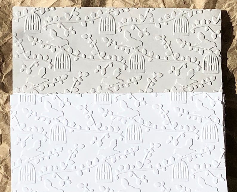 May include: Two sheets of embossed paper with a repeating pattern of birds in cages and branches. The top sheet is a light gray color and the bottom sheet is white.