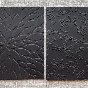K&ouml;nnte beinhalten: Zwei schwarze rechteckige Karten mit gepr&auml;gten Designs. Eine zeigt ein Blumenmuster, die andere ein verziertes, wirbelndes Design. Die Karten sind vor einem strukturierten grauen Hintergrund platziert, der die detaillierten Texturen hervorhebt.