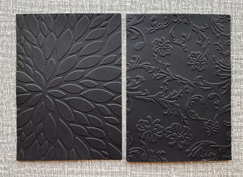 K&ouml;nnte beinhalten: Zwei schwarze rechteckige Karten mit gepr&auml;gten Designs. Eine zeigt ein Blumenmuster, die andere ein verziertes, wirbelndes Design. Die Karten sind vor einem strukturierten grauen Hintergrund platziert, der die detaillierten Texturen hervorhebt.