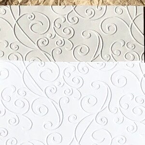 May include: Two sheets of white embossed paper with a swirling pattern. The top sheet is a lighter shade of white.