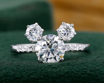 Puede incluir: Un anillo plateado con una piedra central grande, redonda y transparente, flanqueada por dos piedras más pequeñas, que recuerdan a unas orejas de ratón. La banda está adornada con piedras más pequeñas y transparentes. El anillo se encuentra sobre un fondo de terciopelo verde oscuro.