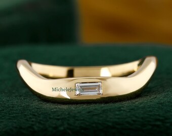 May include: A gold-colored ring with a rectangular, clear gemstone set in the center. The ring has a curved design and is displayed on a dark green surface. The brand name "MicheleJewels" is visible on the ring.