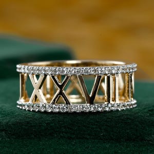 Puede incluir: Un anillo dorado con una banda de pequeñas piedras preciosas transparentes. El anillo presenta números romanos en un diseño recortado. El anillo está sobre un fondo verde oscuro.