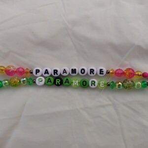 May include: Two beaded bracelets with the word "PARAMORE" spelled out in white letter beads. One bracelet has pink and yellow beads, the other has green beads. Both have colorful accents.