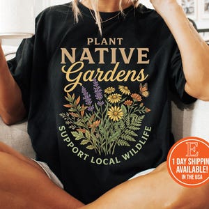 May include: Black t-shirt with the text "PLANT NATIVE Gardens SUPPORT LOCAL WILDLIFE" and a floral design. The shirt has a round neck and short sleeves. The image also shows a white mug.