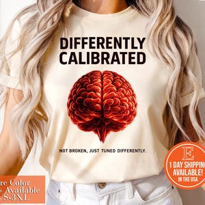 May include: Cream-colored t-shirt featuring the text "DIFFERENTLY CALIBRATED" above a detailed illustration of a red brain. Below the brain, the text reads "NOT BROKEN, JUST TUNED DIFFERENTLY." The shirt is available in sizes S-3XL.