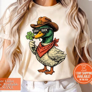 May include: Cream-colored t-shirt featuring a cartoon duck wearing a cowboy hat, sunglasses with "LUCKY DUCK" text, and a red bandana. The duck holds a four-leaf clover. The shirt has the text "+More Color Options Available Sizes S-3XL".