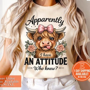 May include: Cream-colored t-shirt featuring a cartoon Highland cow with a pink bow, surrounded by roses. The text reads "Apparently I have an attitude - Who knew?" Sizes S-3XL are available.