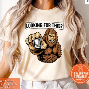 May include: Beige t-shirt with the text "LOOKING FOR THIS?" above a cartoon Bigfoot holding a 10mm object. The Bigfoot is brown and smiling. The shirt has additional text and a logo.
