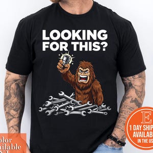 May include: Black t-shirt with the text "LOOKING FOR THIS?" above a cartoon Bigfoot holding a car part, surrounded by wrenches. The shirt is available in sizes S-3XL.
