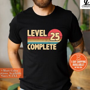May include: Black t-shirt with a retro-style graphic that says "Level 25 Complete" in white text with a red, orange, yellow, and blue color scheme.