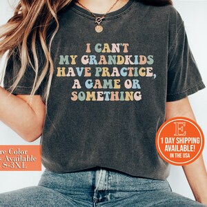 May include: A dark gray t-shirt with the text "I can't my grandkids have practice, a game or something" in colorful letters. The shirt is being worn by a person with long brown hair and blue jeans.