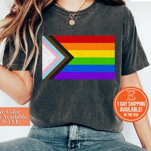 May include: A dark grey t-shirt with a pride flag design featuring a rainbow and a brown, pink, blue, and white triangle. The text "+More Color Options Available Sizes S-3XL" is on the left side of the image.