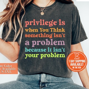May include: A dark gray t-shirt with the text "privilege is when you think something isn't a problem because it isn't your problem" printed in colorful letters.