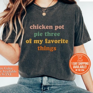 以下が含まれることがあります： カラフルな文字で「チキンポットパイは私の好きなものの3つ」と書かれたグレーのTシャツです