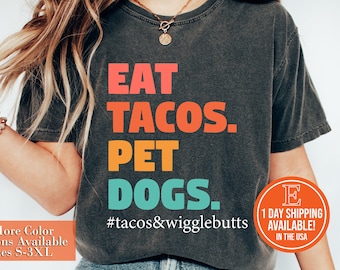 Könnte beinhalten: Ein dunkelgraues T-Shirt mit dem Text "EAT TACOS. PET DOGS." in bunten Buchstaben. Der Text "#tacos&wigglebutts" ist unter dem Haupttext gedruckt.
