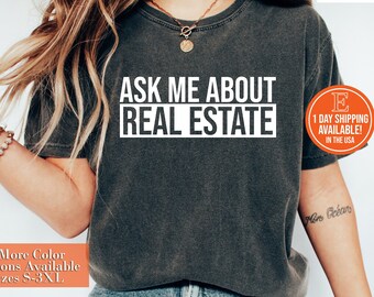 May include: A dark grey t-shirt with the text "ASK ME ABOUT REAL ESTATE" printed in white. The shirt is being worn by a person with long brown hair and blue jeans.