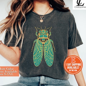 May include: A dark gray t-shirt with a green and yellow cicada graphic. The text "+More Color Options Available Sizes S-3XL" is printed on the bottom left of the shirt.