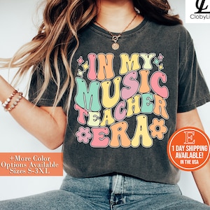 Pode incluir: Uma camiseta cinza escura com um gr&aacute;fico de estilo retr&ocirc; que diz "In My Music Teacher Era" em letras coloridas e contornadas. A camiseta &eacute; usada por uma pessoa com cabelo longo e loiro e &eacute; estilizada com um colar de ouro e uma pulseira de contas.