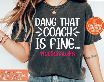 Peut inclure: Un t-shirt gris avec le texte "DANG THAT COACH IS FINE... #coachswife" imprimé en blanc. Le texte "#coachswife" est en rose.