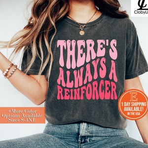 May include: A gray t-shirt with the text "THERE'S ALWAYS A REINFORCER" in pink and white lettering. The shirt is being worn by a person with long brown hair.