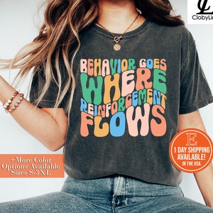 May include: A dark gray t-shirt with the text "BEHAVIOR GOES WHERE REINFORCEMENT FLOWS" in colorful, wavy letters.