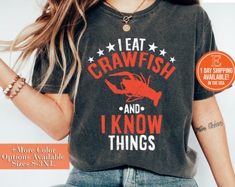 May include: A dark gray t-shirt with a white graphic that says "I eat crawfish and I know things". The graphic also includes a red crawfish illustration. The shirt is being worn by a person with long brown hair.