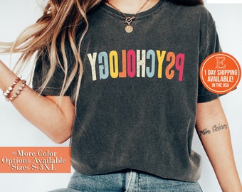May include: A dark grey t-shirt with the word "Psychology" printed in rainbow colors. The word is stylized with each letter in a different color. The shirt is being worn by a person with long brown hair.