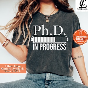 May include: A dark gray t-shirt with the text "Ph.D. IN PROGRESS" and a progress bar graphic. The text is in white. The shirt is being worn by a person.