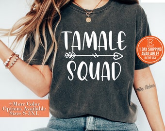 以下が含まれることがあります： 白い文字で「TAMALE SQUAD」と書かれた濃いグレーのTシャツ。テキストには、ハートの付いた白い矢印が添えられています。このTシャツは、長い茶色の髪をした人が着用しています。