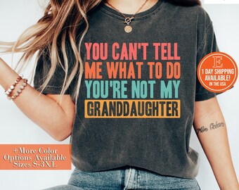 May include: A dark gray t-shirt with the text "YOU CAN'T TELL ME WHAT TO DO YOU'RE NOT MY GRANDDAUGHTER" in colorful letters. The shirt is being worn by a person with long blonde hair.