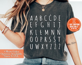 Op de afbeelding: Een donkergrijs T-shirt met een witte afbeelding van het alfabet, inclusief accenten. De letters zijn gerangschikt in een raster. Het shirt wordt gedragen door een persoon met lang bruin haar.
