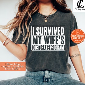 May include: A dark grey t-shirt with the text "I Survived My Wife's Doctorate Program" printed in white. The text has a distressed, vintage look.