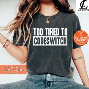 May include: A dark gray t-shirt with the text "TOO TIRED TO CODESWITCH" in white letters. The shirt is being worn by a person with long brown hair and blue jeans.