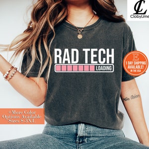 May include: Dark gray t-shirt with the text "RAD TECH LOADING" in white, above a pink loading bar. The shirt also has the text "+More Color Options Available Sizes S-3XL".