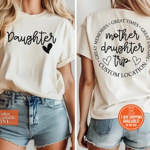 Peut inclure: T-shirt crème avec le mot "Daughter" en écriture cursive et un cœur noir. Le dos du t-shirt présente un motif circulaire avec le texte "Mother Daughter Trip" et "Custom Location". Disponible en tailles S-3XL.