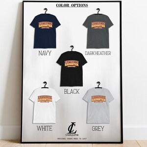 Puede incluir: Una exhibici&oacute;n enmarcada de camisetas en varios colores: azul marino, gris oscuro, negro, blanco y gris. Cada camiseta presenta el texto "FAVORITE DAUGHTER SINCE 1999" en un estilo retro. La exhibici&oacute;n incluye el logotipo de la marca.