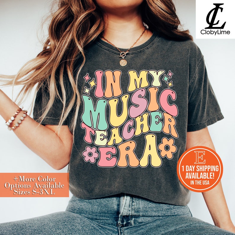 Pode incluir: Uma camiseta cinza escura com um gr&aacute;fico de estilo retr&ocirc; que diz "In My Music Teacher Era" em letras coloridas e contornadas. A camiseta &eacute; usada por uma pessoa com cabelo longo e loiro e &eacute; estilizada com um colar de ouro e uma pulseira de contas.