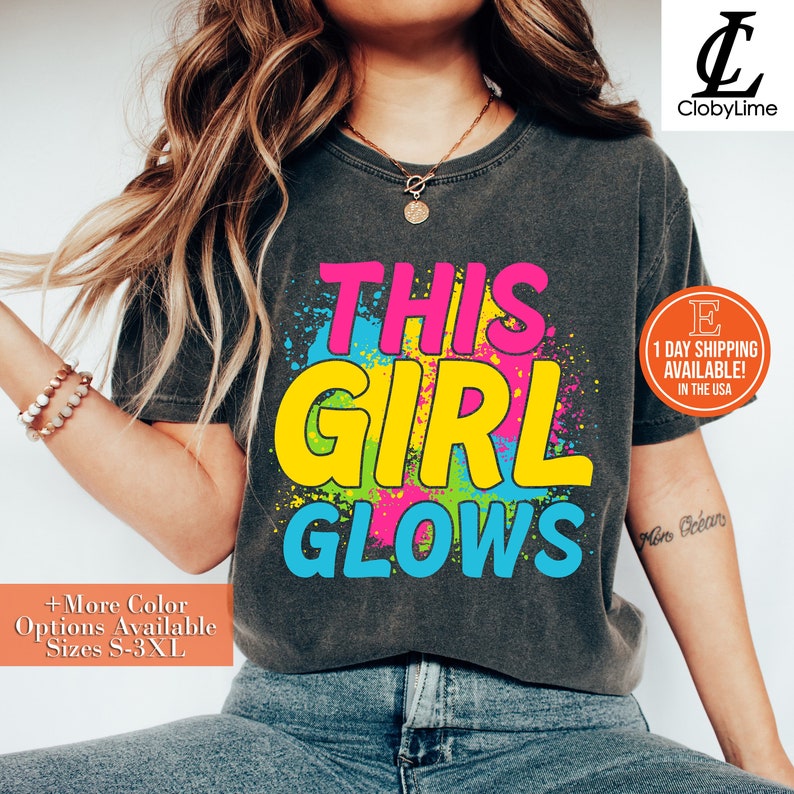 Puede incluir: Una camiseta gris con el texto "THIS GIRL GLOWS" en letras de colores. Las letras est&aacute;n delineadas en negro y tienen un efecto de salpicadura de pintura.