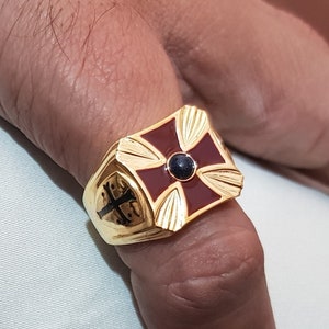 May include: A gold ring featuring a red enamel cross with a black stone at its centre. The ring is worn on a finger.