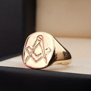 Può includere: Un anello a sigillo in oro con un designo a quadrato e compasso. L'anello è in una scatola per anelli nera.