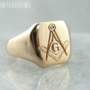May include: A gold signet ring featuring an engraved Masonic symbol on the top. The symbol is a compass and square with a 'G' at its centre.