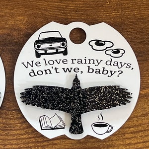 May include: White oval-shaped tag with black text and images. The text reads "We love rainy days, don't we, baby?"  The images include a car, a book, a cup of coffee, and a black crow with glitter.