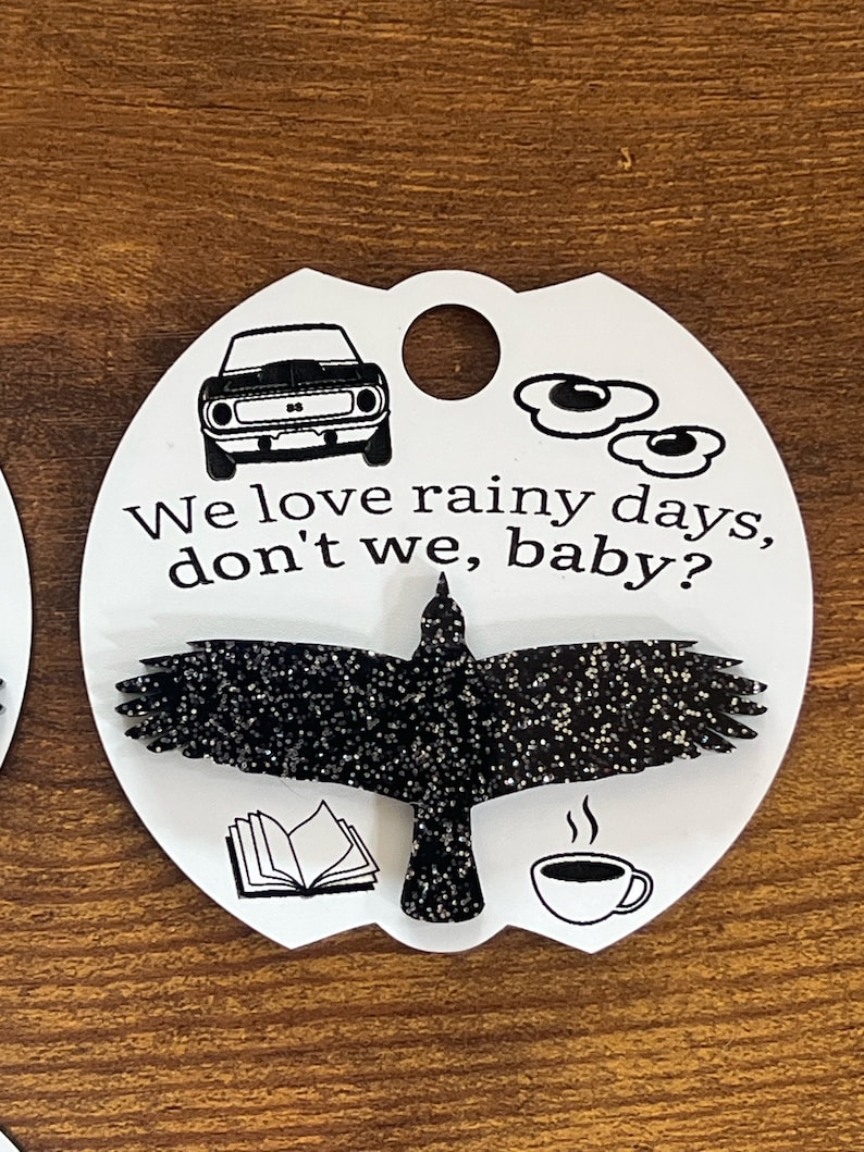 May include: White oval-shaped tag with black text and images. The text reads "We love rainy days, don't we, baby?"  The images include a car, a book, a cup of coffee, and a black crow with glitter.