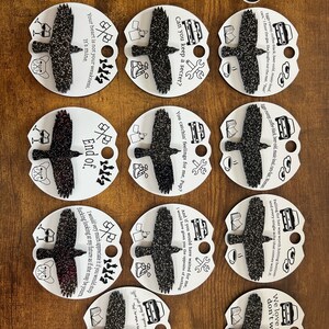 May include: Nine round white and black paper ornaments with a black crow design and text. The text includes quotes such as "Your heart is not your weakness, it's mine." and "Can you keep a secret?"